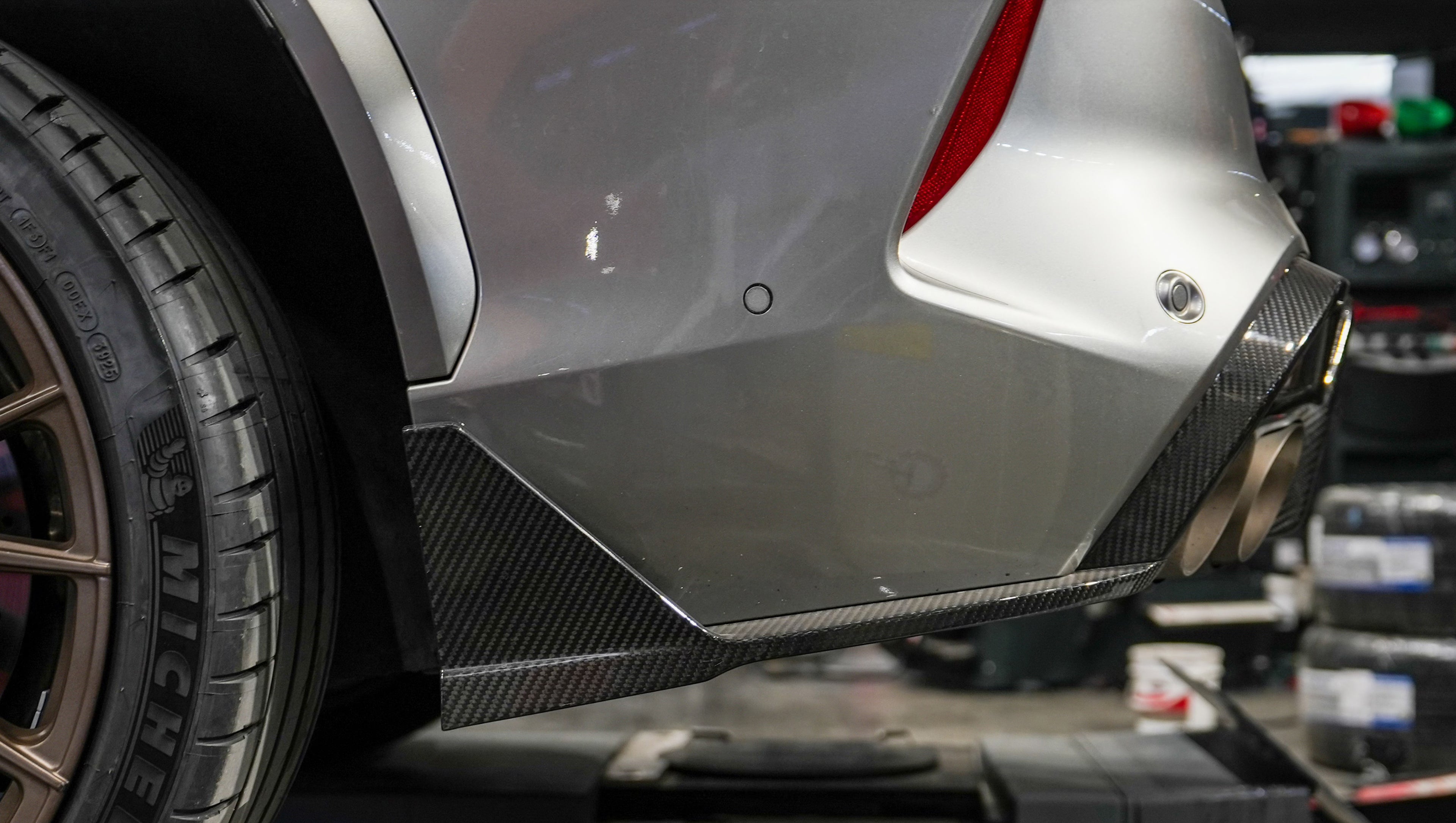 Gray BMW X5M featuring a REVOZPORT carbon fiber body kit with aggressive front lip, enlarged air intakes, side skirts, and sporty aero styling displayed inside a performance workshop.