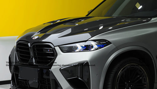 Front view of a white BMW X5M featuring a REVOZPORT carbon fiber hood, aggressive front bumper, enlarged air intakes, and carbon fiber front lip displayed inside a modern showroom.