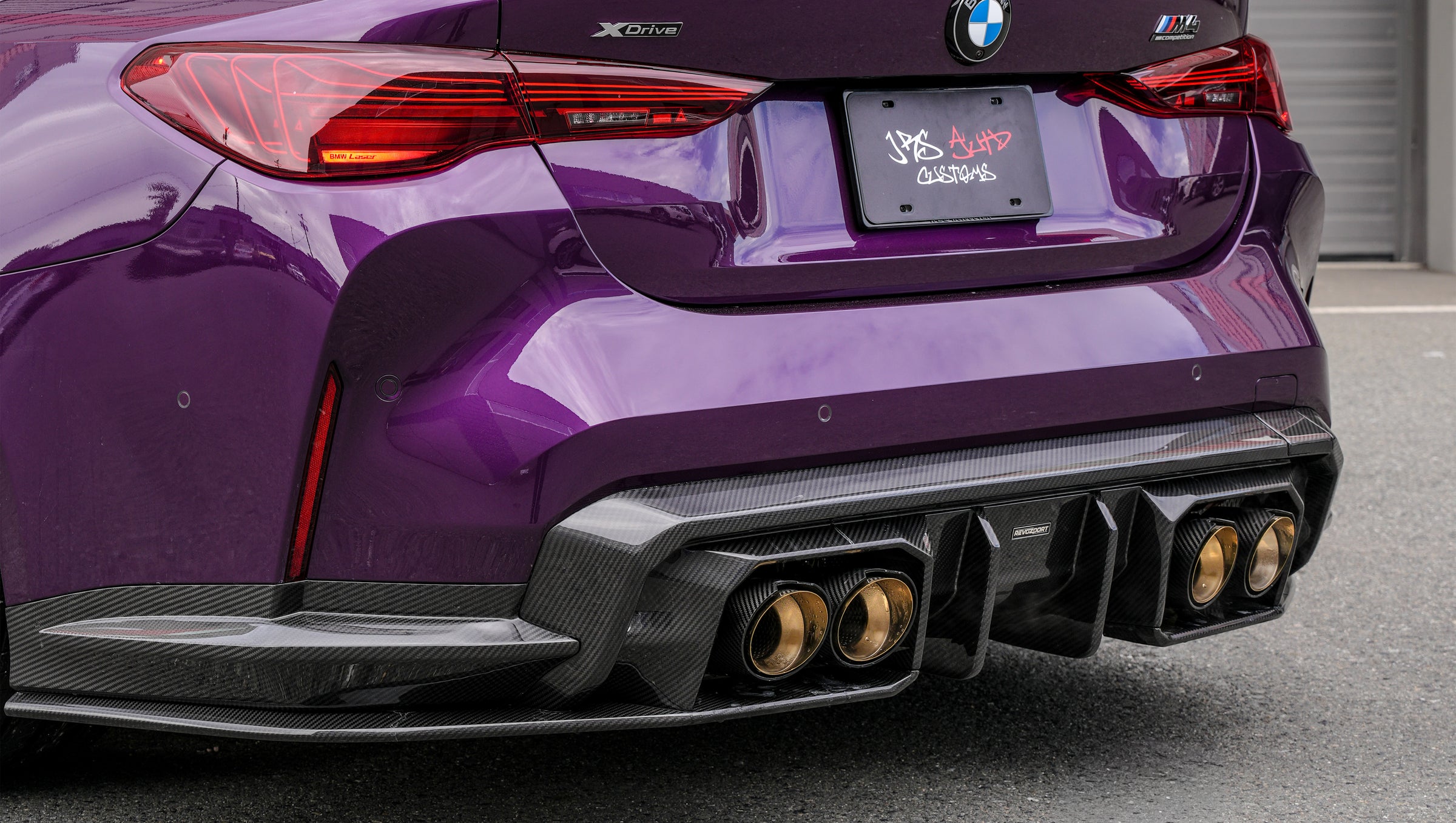 Purple BMW G82 M4 featuring a REVOZPORT carbon fiber body kit with front lip, side skirts, vented fenders, and gold wheels parked outside a modern industrial building.