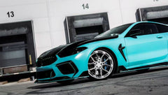Turquoise BMW F92 M8 featuring a REVOZPORT carbon fiber body kit with front lip, side skirts, black hood accents, side vents, and silver forged wheels parked outside an industrial loading dock.