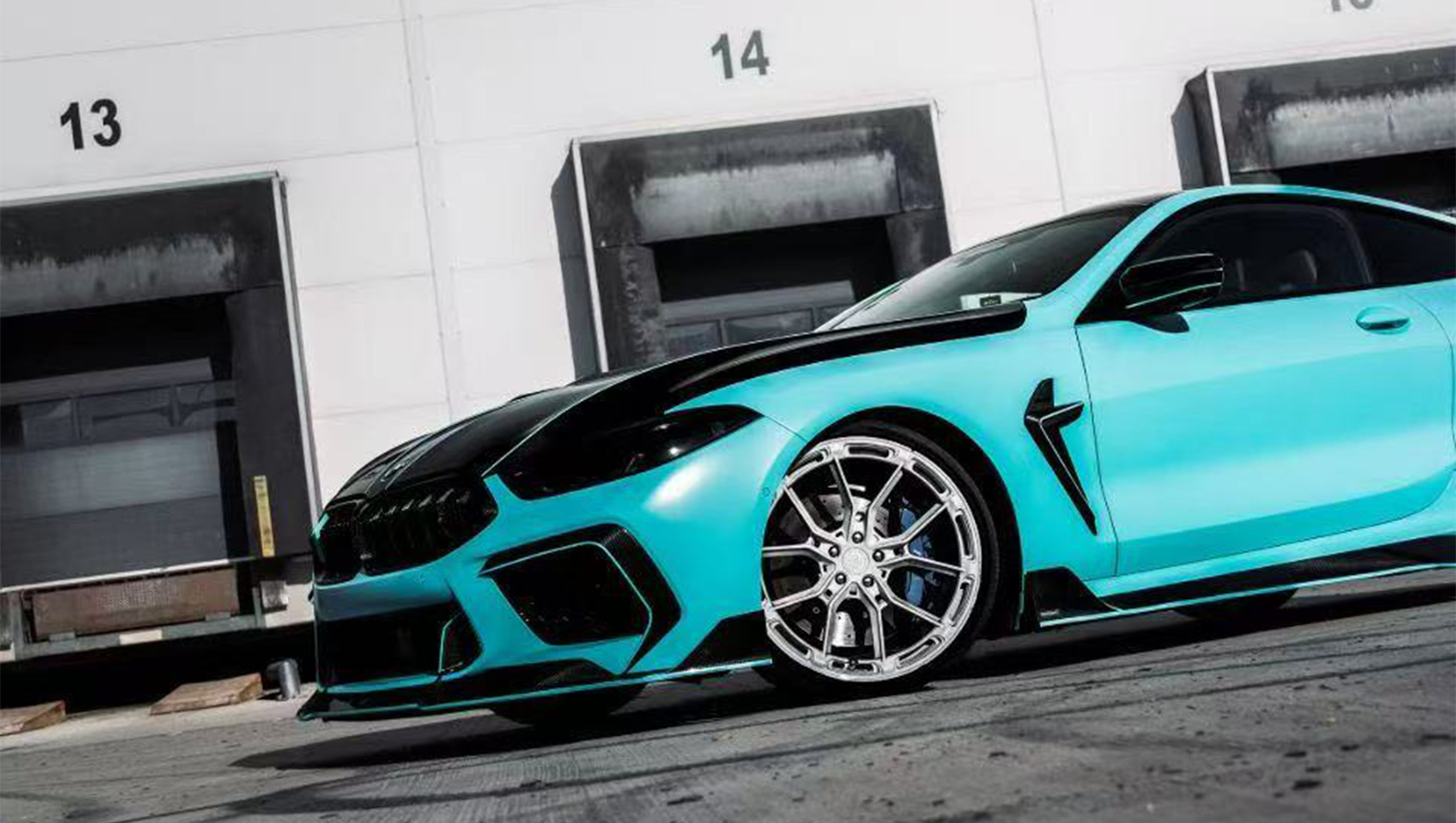 Turquoise BMW F92 M8 featuring a REVOZPORT carbon fiber body kit with front lip, side skirts, black hood accents, side vents, and silver forged wheels parked outside an industrial loading dock.