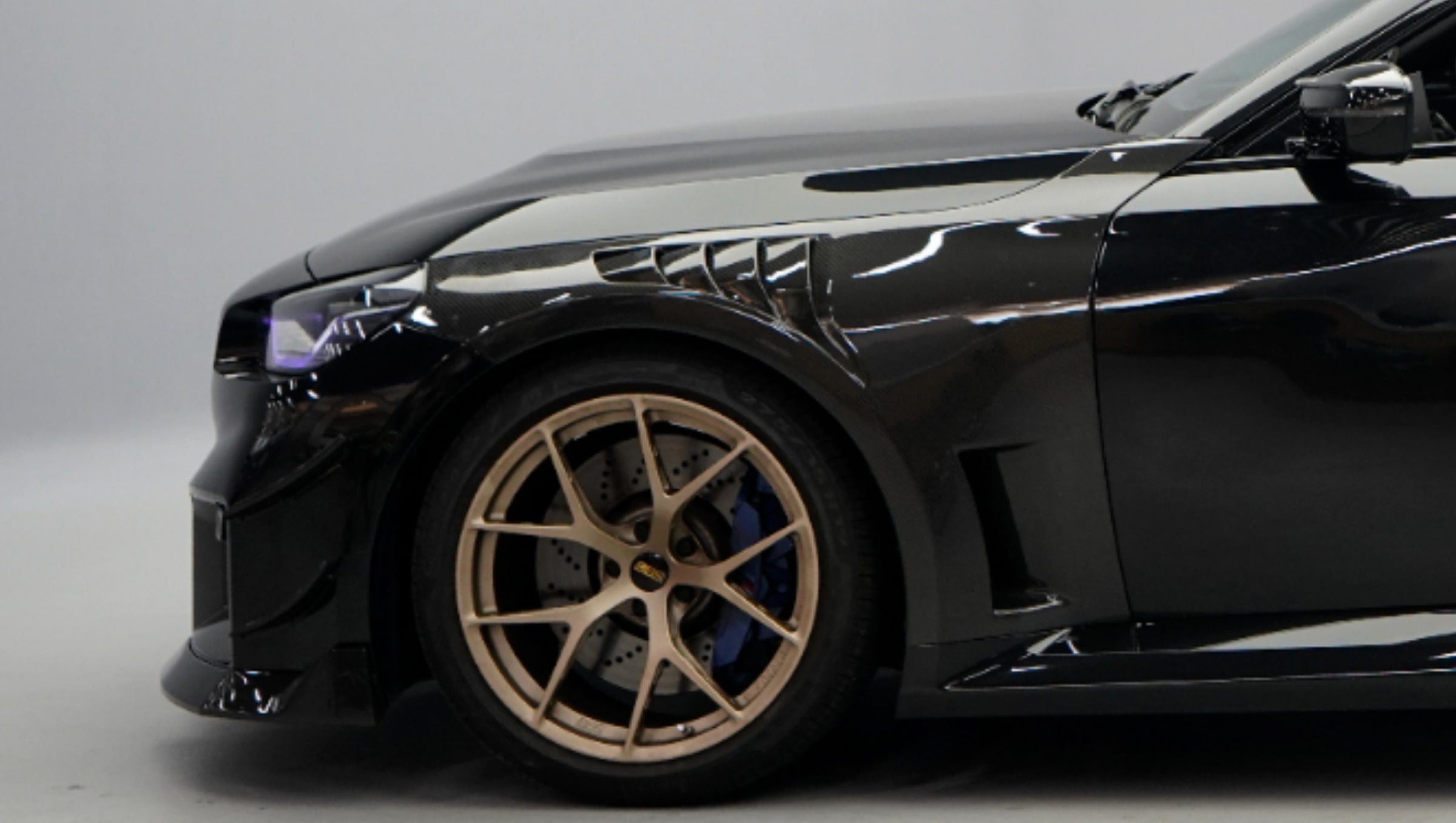 Front close-up of a black BMW G87 M2 featuring an aggressive carbon fiber front bumper, deep front splitter, large air intakes, and gloss carbon fiber aero details in a studio setting.