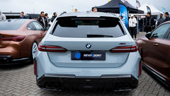 Gray BMW G90 M5 Touring featuring a REVOZPORT carbon fiber front lip, aggressive front aero styling, and black grille displayed at a car event.