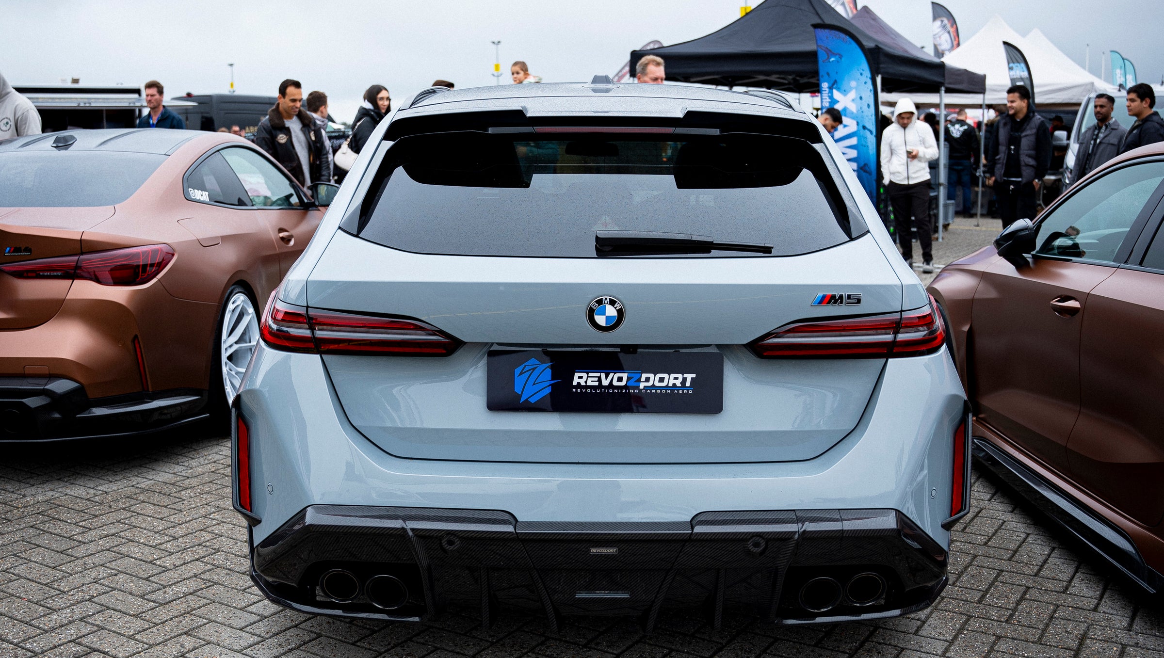 Gray BMW G90 M5 Touring featuring a REVOZPORT carbon fiber front lip, aggressive front aero styling, and black grille displayed at a car event.