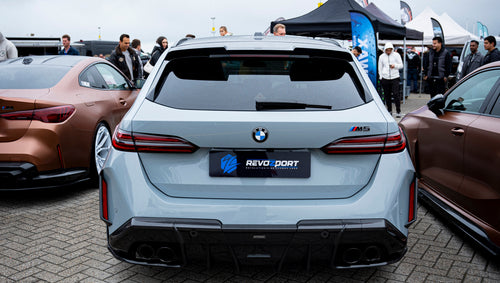 Gray BMW G90 M5 Touring featuring a REVOZPORT carbon fiber front lip, aggressive front aero styling, and black grille displayed at a car event.