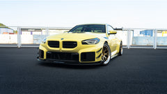 Pastel yellow BMW G82 M4 with carbon fiber front splitter, hood vents, side canards, bronze wheels, and large rear wing parked at a race track facility during golden hour.