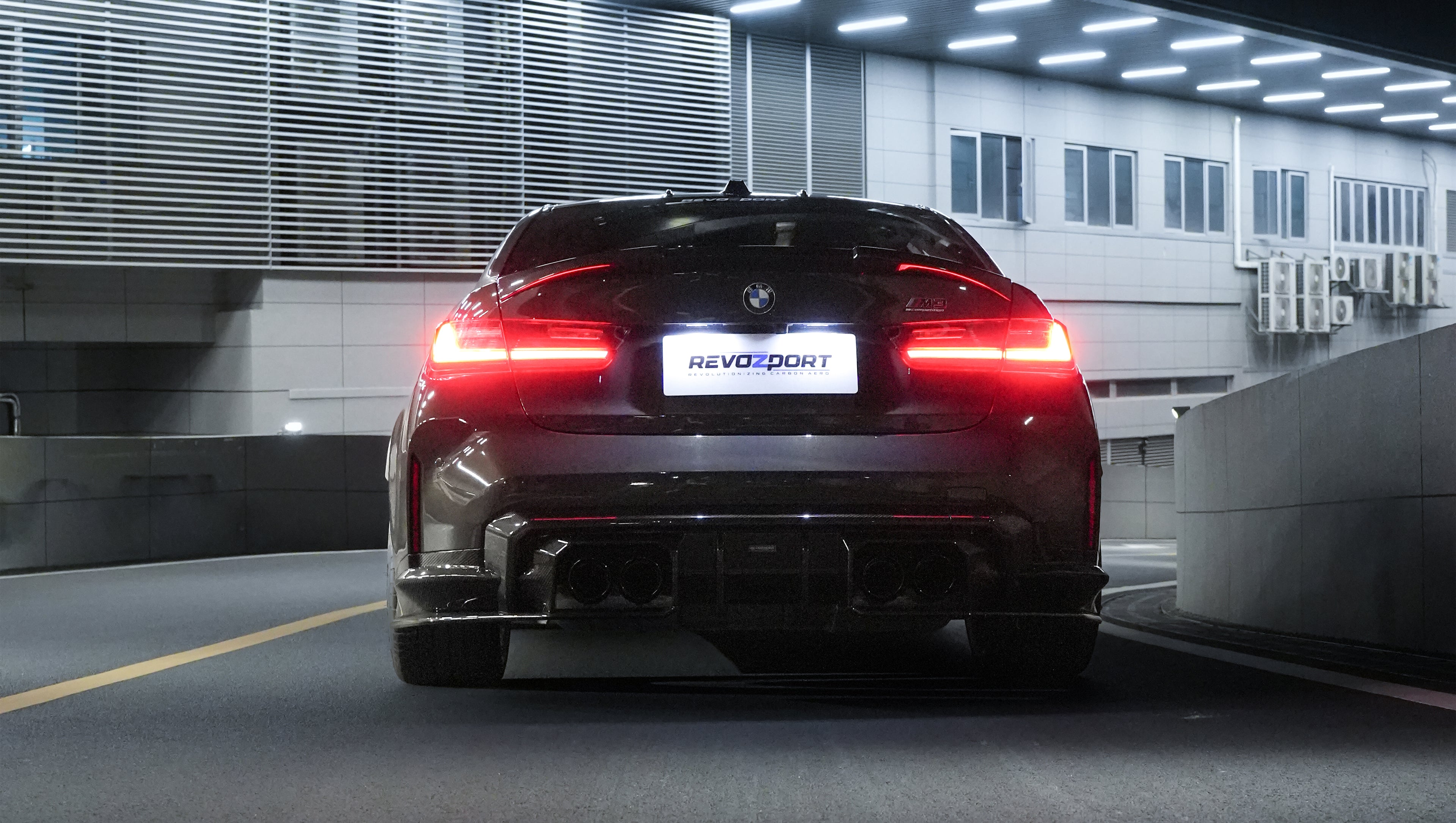 Gray BMW G80 M3 featuring a REVOZPORT carbon fiber front lip, side skirts, bronze wheels, and M The M3 side graphics driving through a tunnel at night.