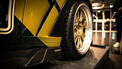 BMW G80 M3 race build with REVOZPORT carbon fiber aero kit, gold wheels, and black-yellow motorsport livery displayed at an indoor automotive exhibition.