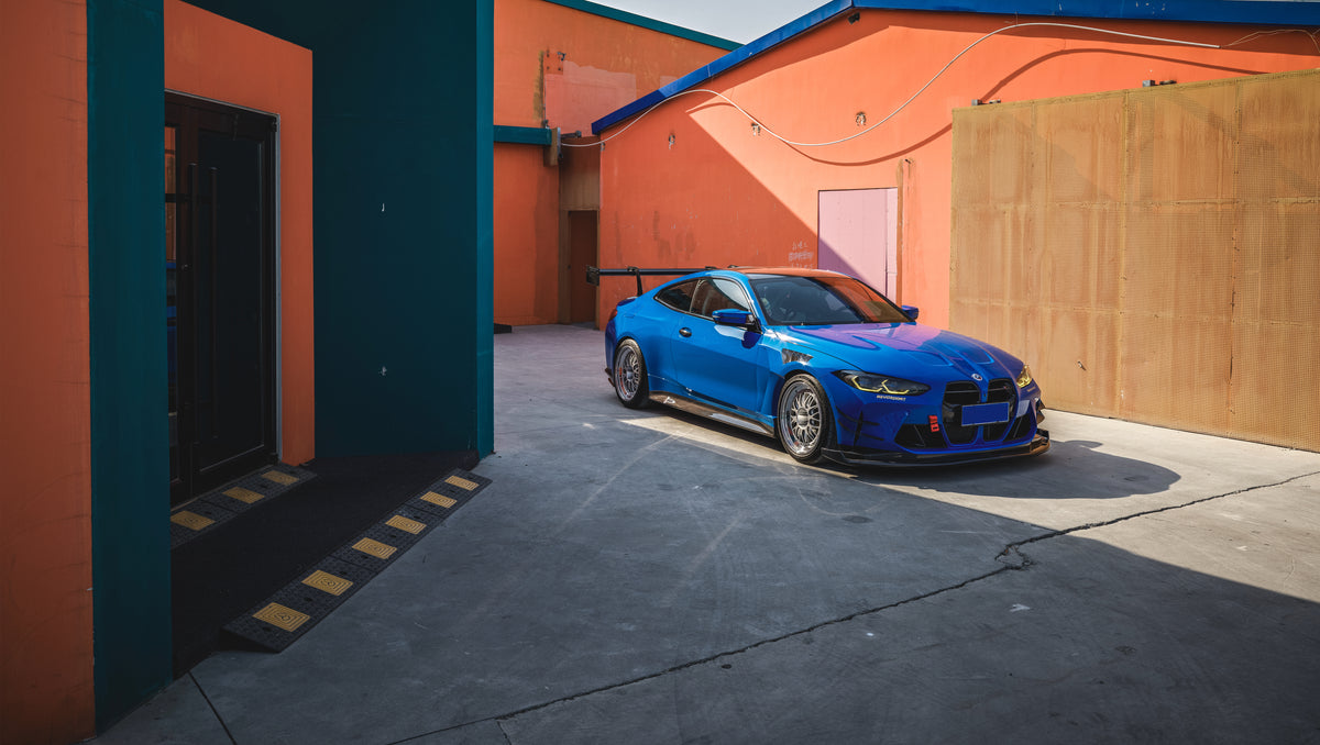 Blue BMW G82 M4 with carbon fiber front splitter, side skirts, rear wing, and aftermarket wheels parked in a modern industrial courtyard with colorful architectural walls.