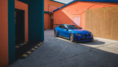 Blue BMW G82 M4 with carbon fiber front splitter, side skirts, rear wing, and aftermarket wheels parked in a modern industrial courtyard with colorful architectural walls.