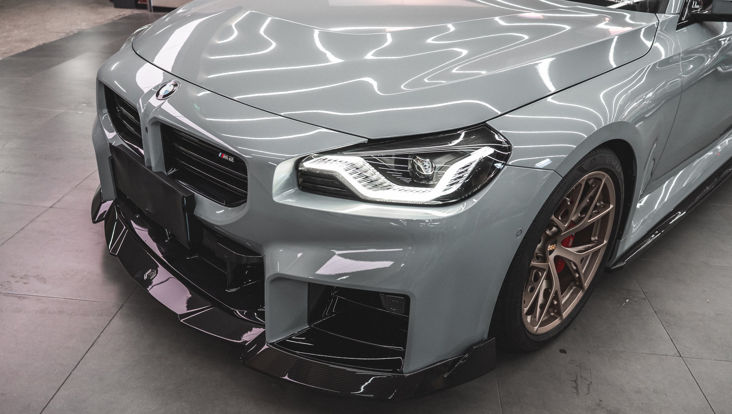 Gray BMW G87 M2 featuring a REVOZPORT carbon fiber front lip, aggressive front aero styling, bronze wheels, and red performance brakes displayed in a modern indoor garage.