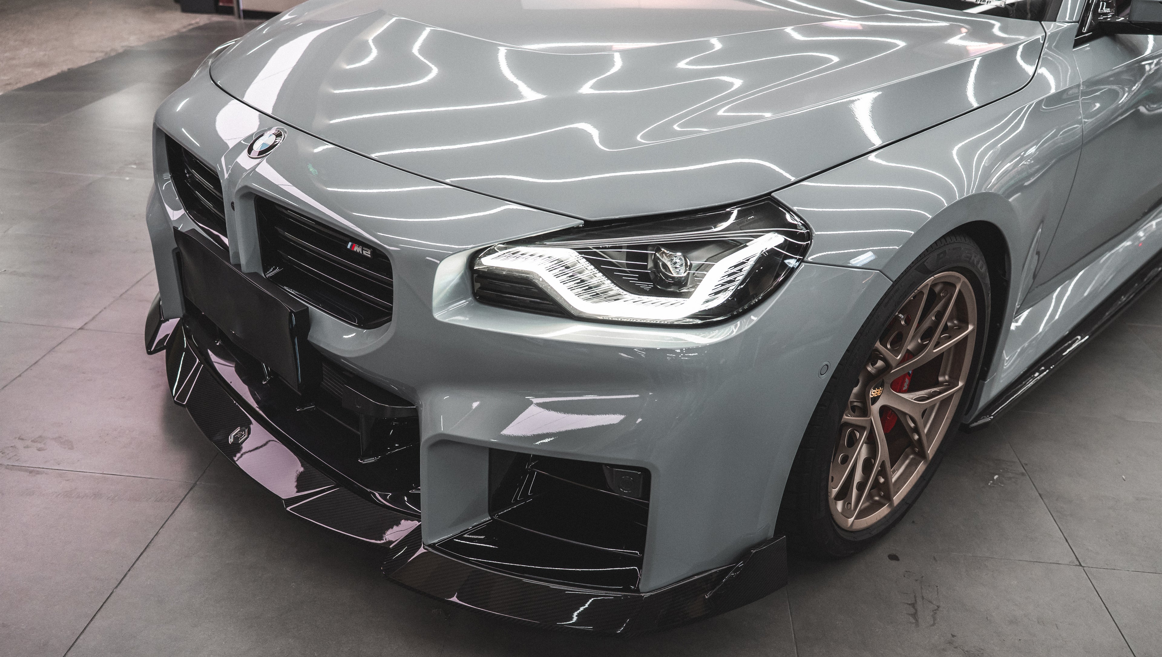 Gray BMW G87 M2 featuring a REVOZPORT carbon fiber front lip, aggressive front aero styling, bronze wheels, and red performance brakes displayed in a modern indoor garage.