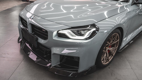 Gray BMW G87 M2 featuring a REVOZPORT carbon fiber front lip, aggressive front aero styling, bronze wheels, and red performance brakes displayed in a modern indoor garage.