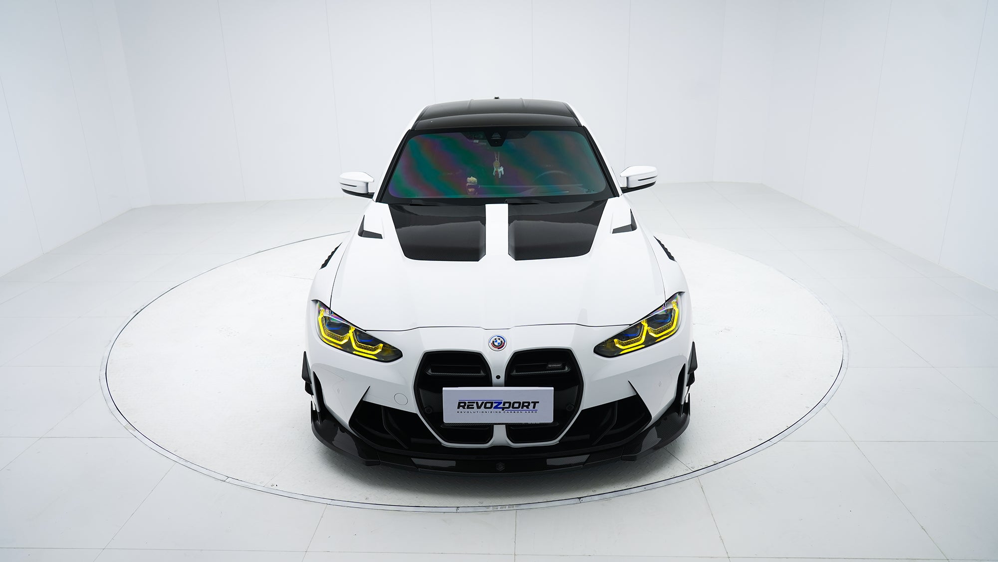 White BMW G80 M3 featuring a REVOZPORT carbon fiber body kit with front lip, side skirts, canards, vented fenders, and yellow DRL headlights displayed in a clean studio setting.
