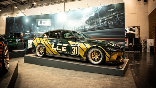 BMW G80 M3 race build with REVOZPORT carbon fiber aero kit, gold wheels, and black-yellow motorsport livery displayed at an indoor automotive exhibition.