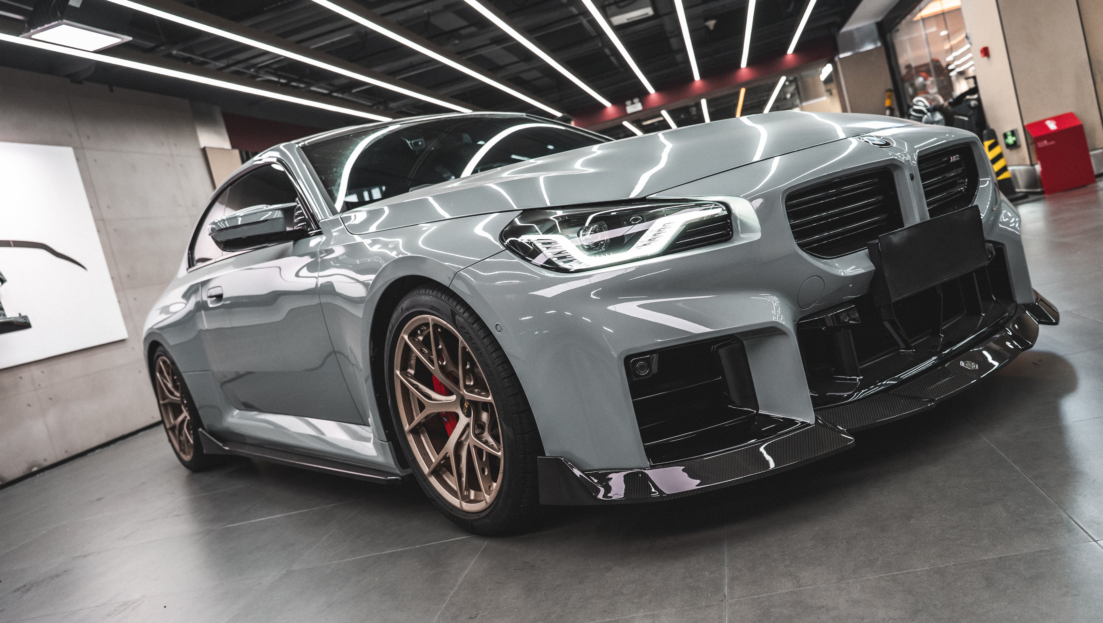 Gray BMW G87 M2 featuring a REVOZPORT carbon fiber front lip, aggressive front aero styling, bronze wheels, and red performance brakes displayed in a modern indoor garage.