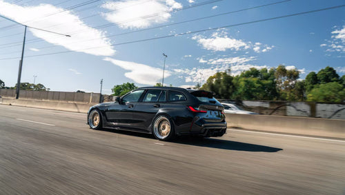 Black BMW G81 M3 Touring featuring a REVOZPORT carbon fiber body kit with vented fenders, side skirts, bronze wheels, and red performance brakes parked beside a stone wall.