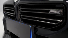 Front close-up of a black BMW G87 M2 featuring an aggressive carbon fiber front bumper, deep front splitter, large air intakes, and gloss carbon fiber aero details in a studio setting.