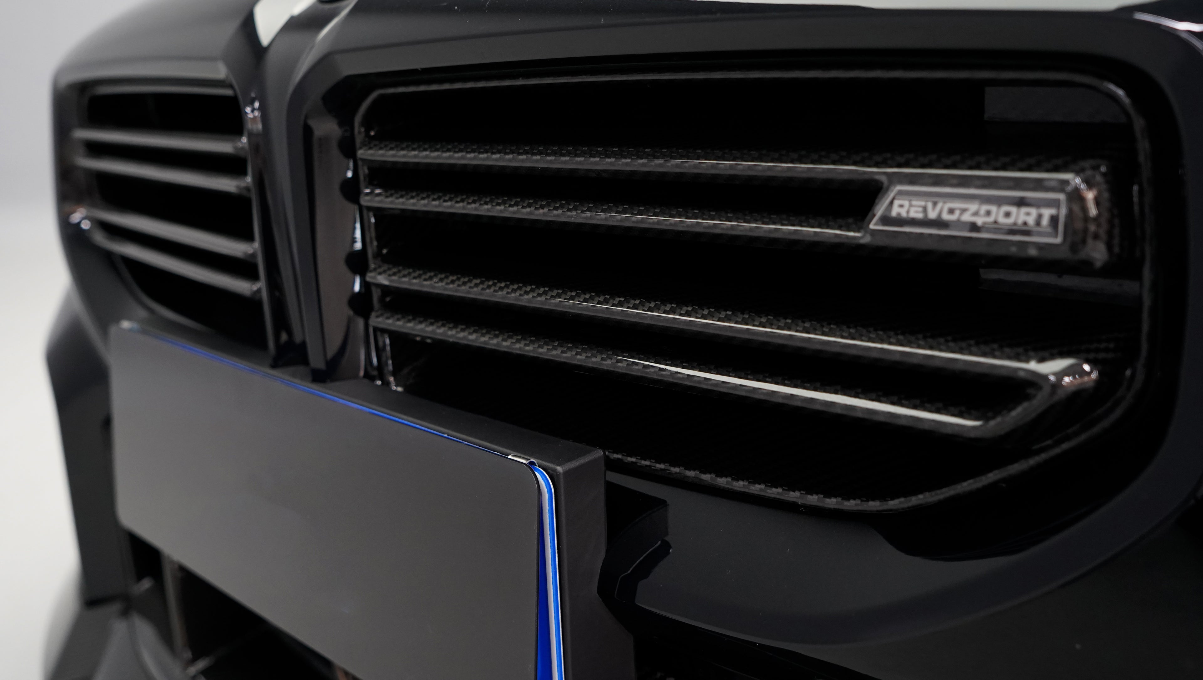 Front close-up of a black BMW G87 M2 featuring an aggressive carbon fiber front bumper, deep front splitter, large air intakes, and gloss carbon fiber aero details in a studio setting.