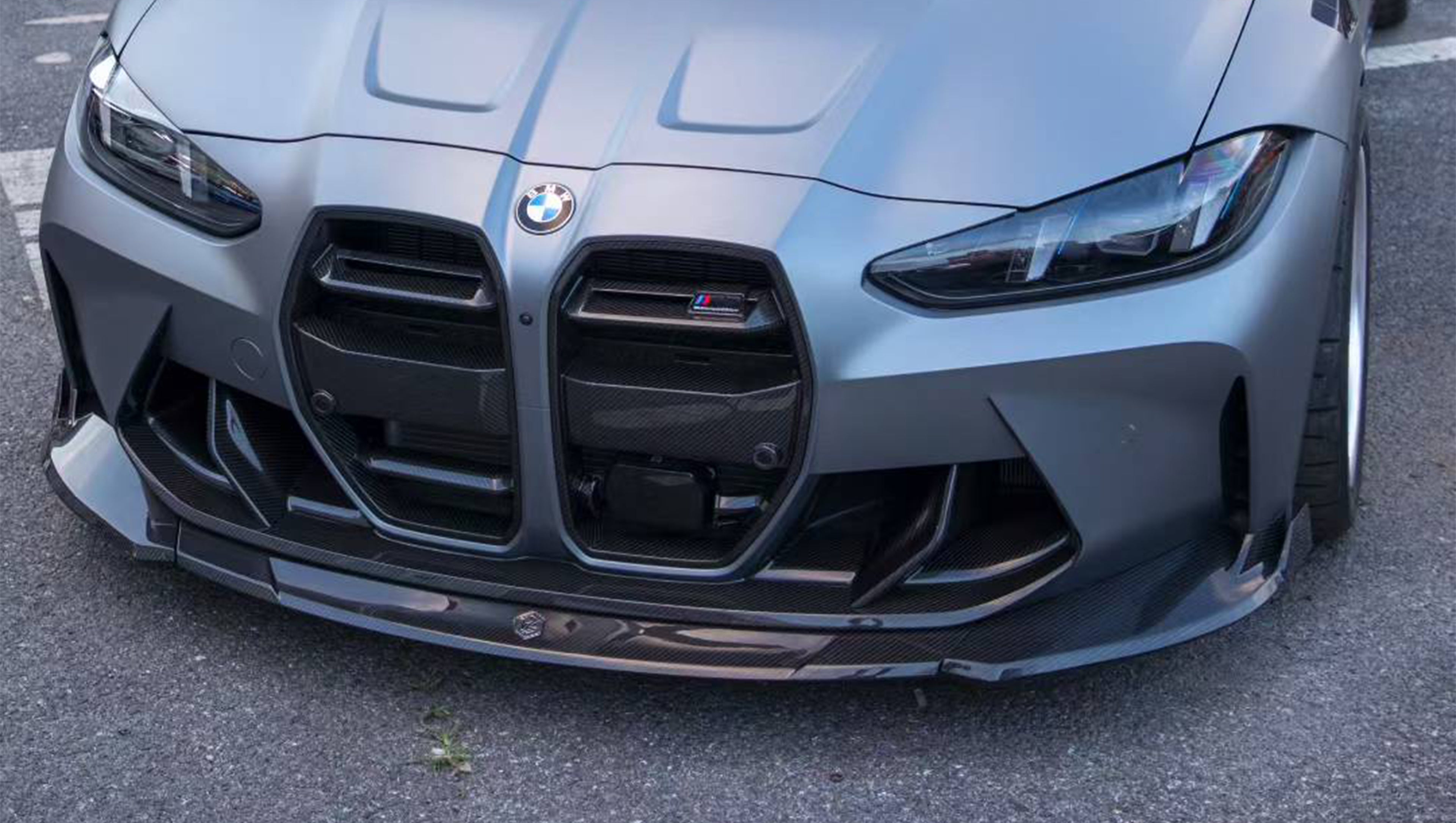 Close-up of a gray BMW G80 M3 featuring a REVOZPORT carbon fiber front lip, vented fenders, side skirts, and aggressive front aero styling in an outdoor parking lot.