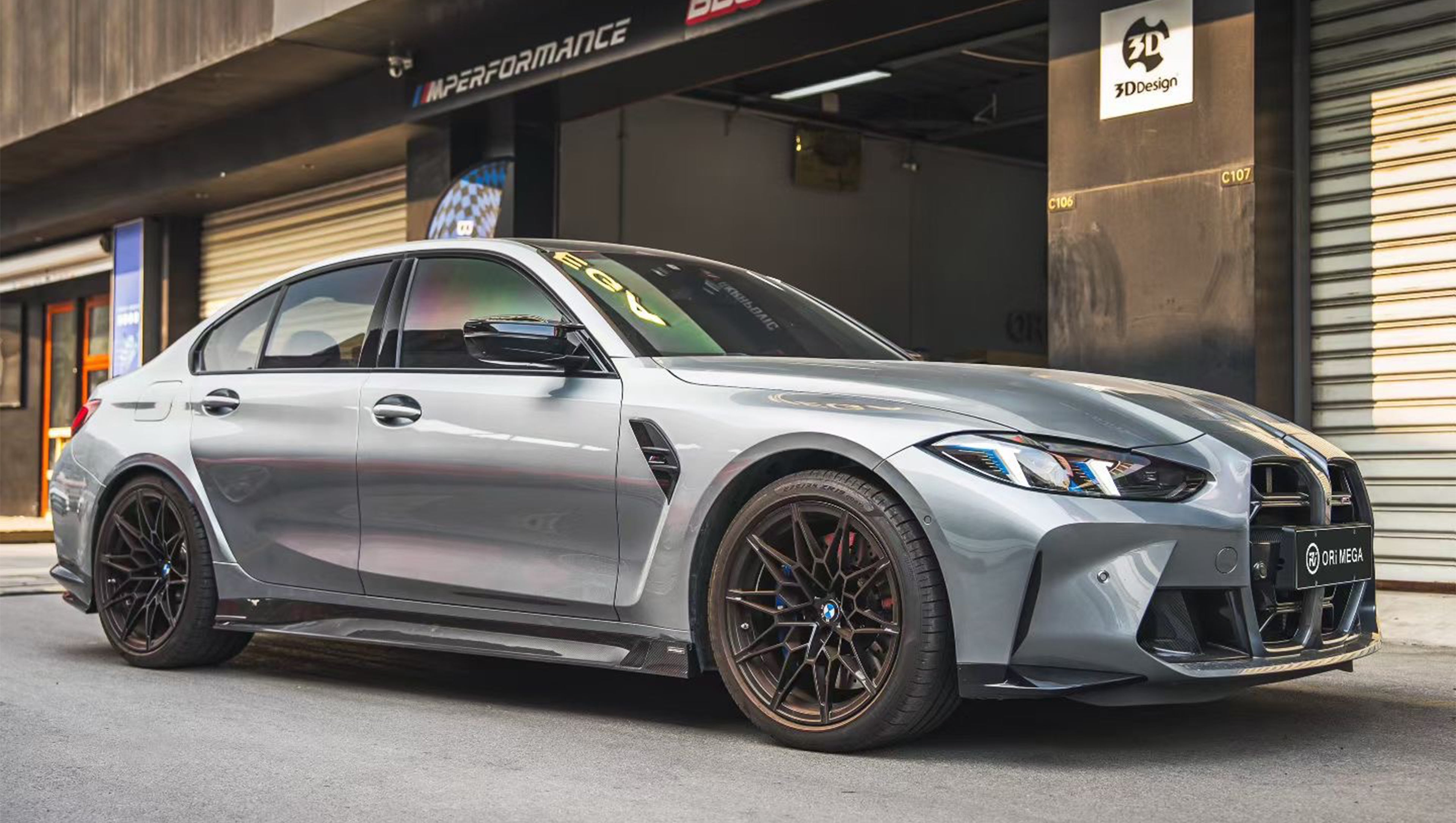 Gray BMW G80 M3 featuring a REVOZPORT carbon fiber front lip, side skirts, and aggressive aero styling parked outside a modern urban garage.
