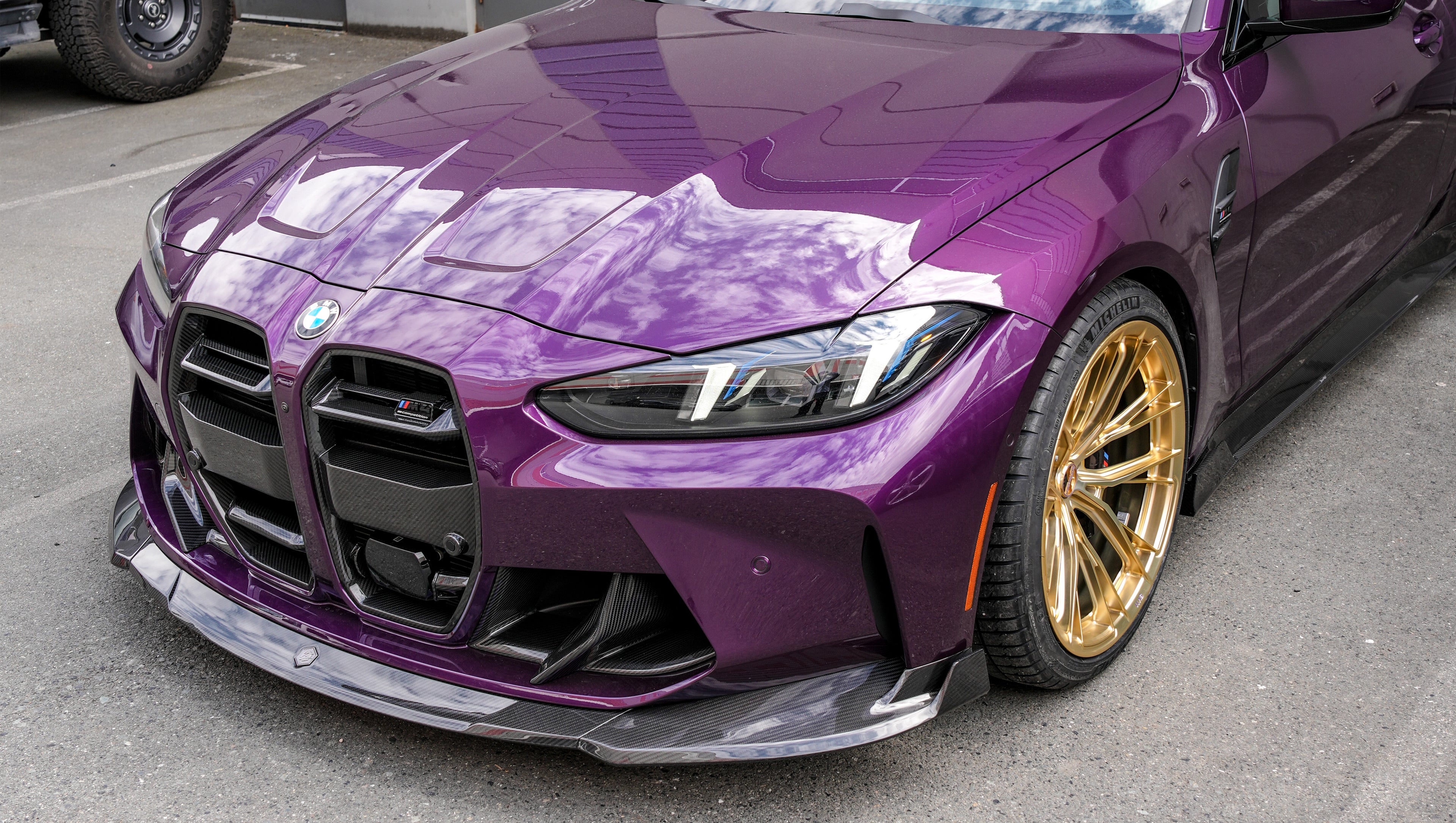 Purple BMW G82 M4 featuring a REVOZPORT carbon fiber body kit with front lip, side skirts, vented fenders, and gold wheels parked outside a modern industrial building.