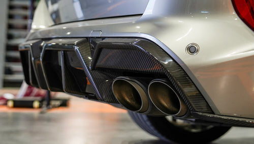 Gray BMW X5M featuring a REVOZPORT carbon fiber body kit with aggressive front lip, enlarged air intakes, side skirts, and sporty aero styling displayed inside a performance workshop.