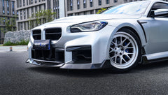 Close-up of a silver BMW G87 M2 featuring a REVOZPORT carbon fiber front lip, aggressive front aero styling, side skirt details, and white multi-spoke wheels parked in a modern urban setting.