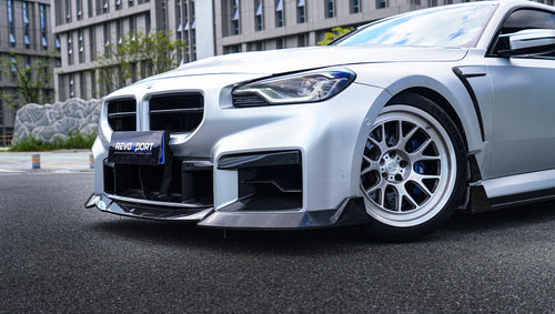 Close-up of a silver BMW G87 M2 featuring a REVOZPORT carbon fiber front lip, aggressive front aero styling, side skirt details, and white multi-spoke wheels parked in a modern urban setting.