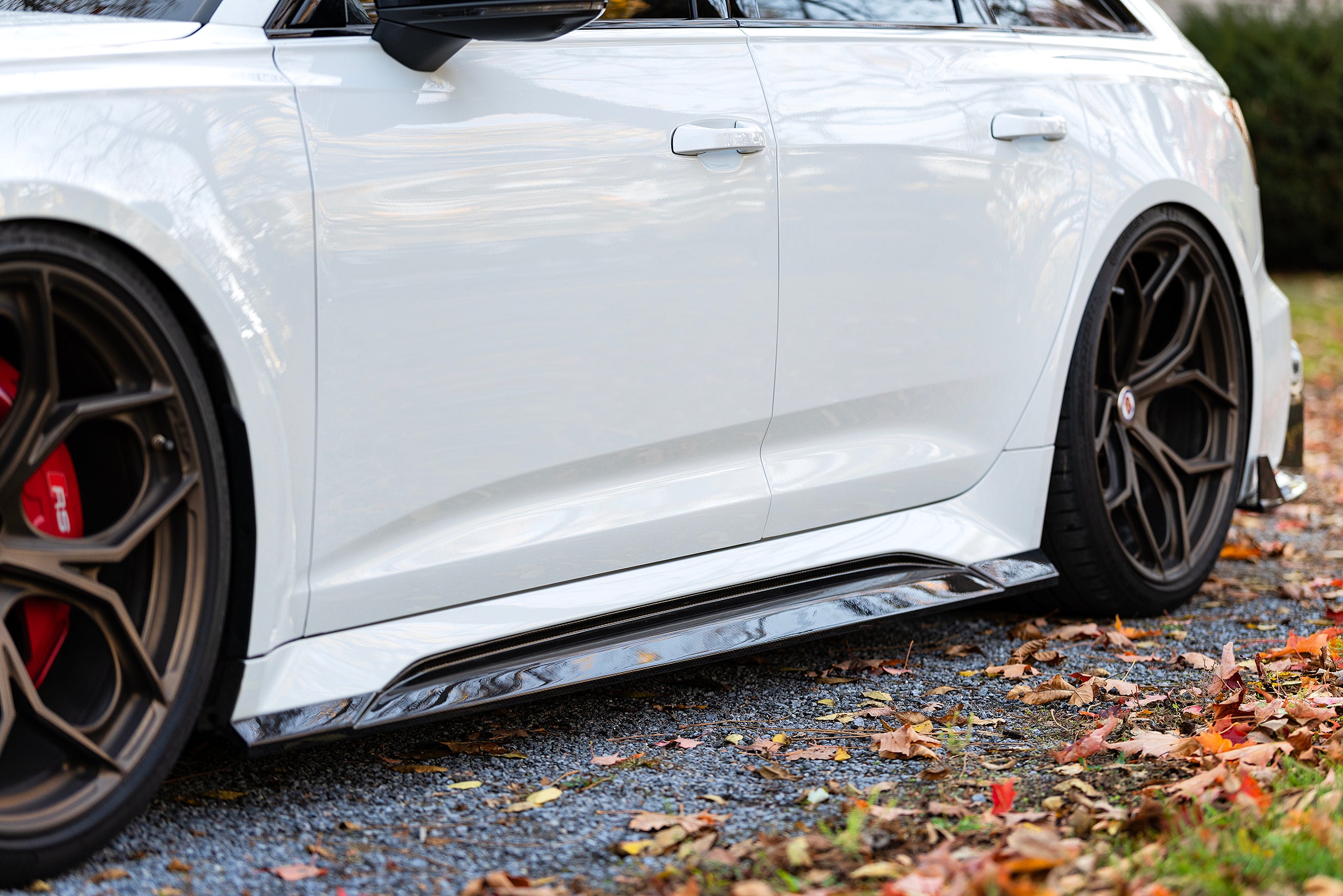White Audi RS6 Avant featuring a REVOZPORT carbon fiber body kit with front lip, side skirts, and aggressive aero styling parked in front of a historic stone mansion during autumn.