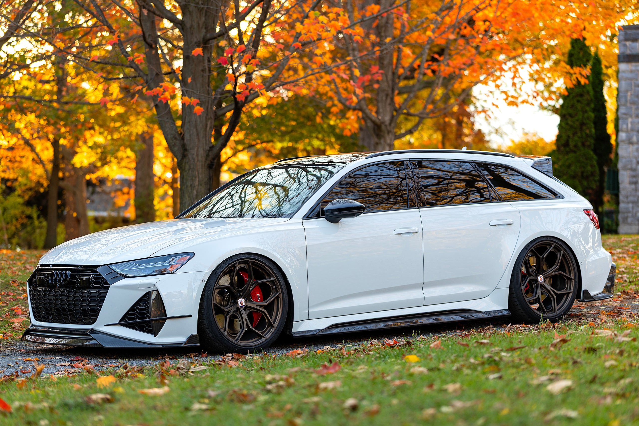 White Audi RS6 Avant featuring a REVOZPORT carbon fiber body kit with front lip, side skirts, and aggressive aero styling parked in front of a historic stone mansion during autumn.