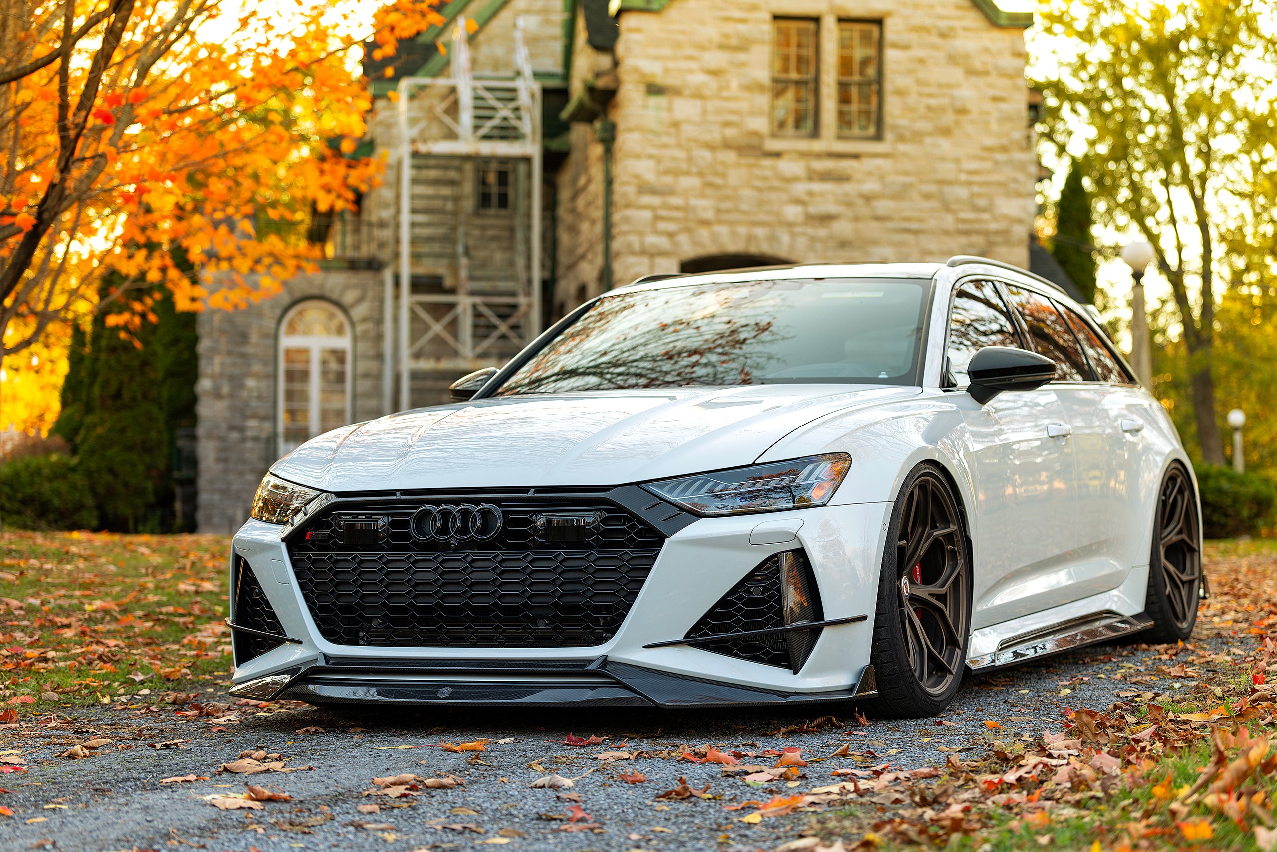 White Audi RS6 Avant featuring a REVOZPORT carbon fiber body kit with front lip, side skirts, and aggressive aero styling parked in front of a historic stone mansion during autumn.