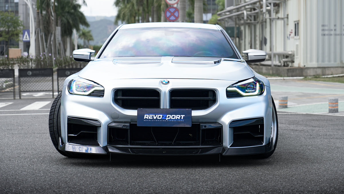 Front view of a silver BMW G87 M2 featuring a REVOZPORT carbon fiber front lip, aggressive front bumper aero styling, and vented hood parked on an urban street.