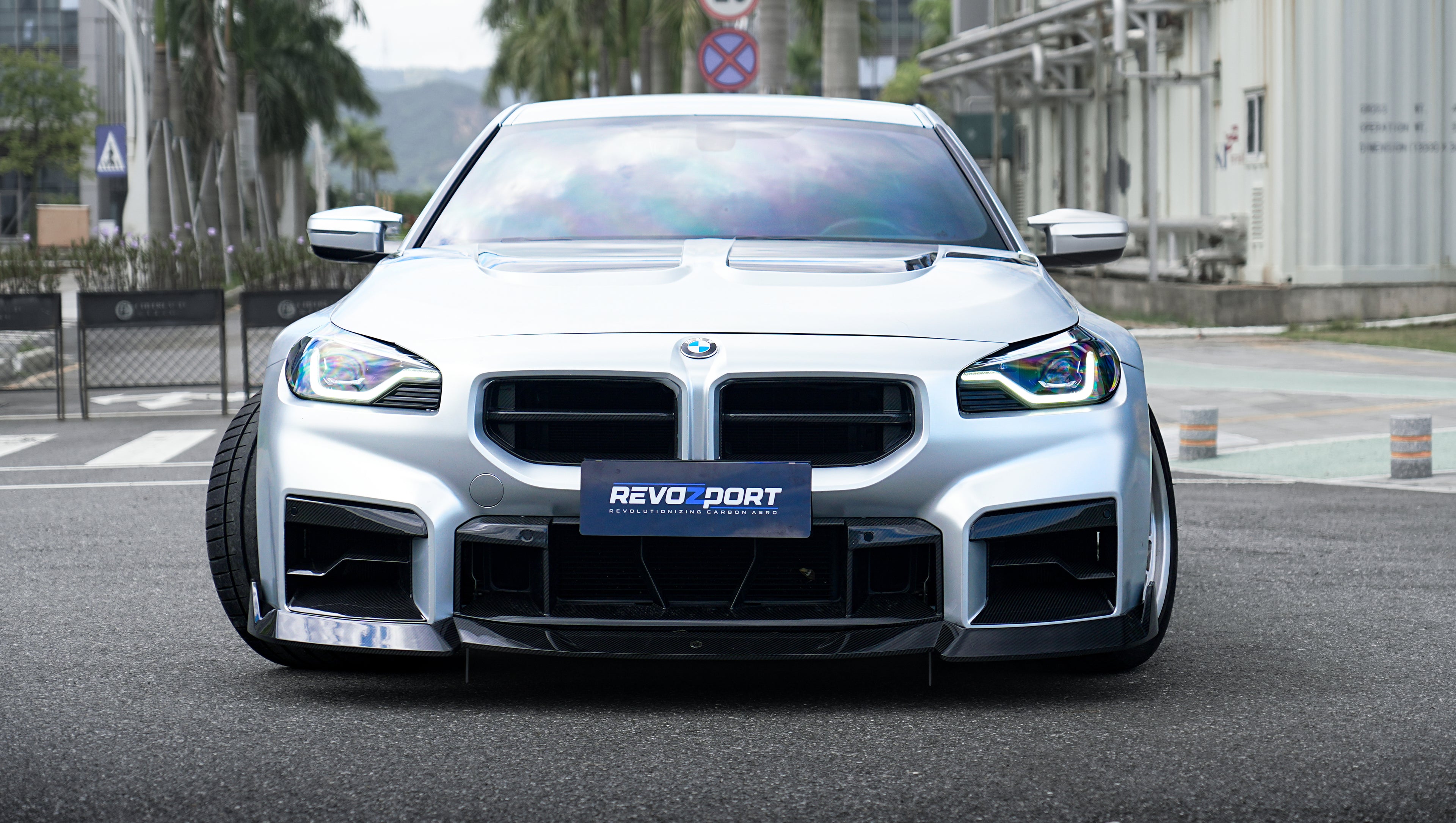 Front view of a silver BMW G87 M2 featuring a REVOZPORT carbon fiber front lip, aggressive front bumper aero styling, and vented hood parked on an urban street.