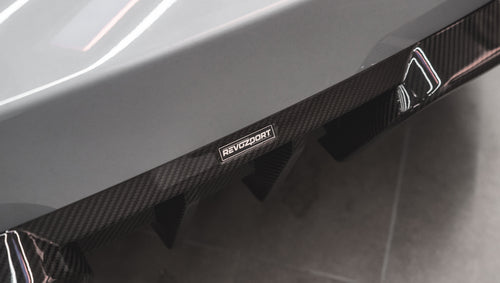 Gray BMW G87 M2 featuring a REVOZPORT carbon fiber front lip, aggressive front aero styling, bronze wheels, and red performance brakes displayed in a modern indoor garage.