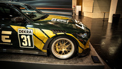 BMW G80 M3 race build with REVOZPORT carbon fiber aero kit, gold wheels, and black-yellow motorsport livery displayed at an indoor automotive exhibition.