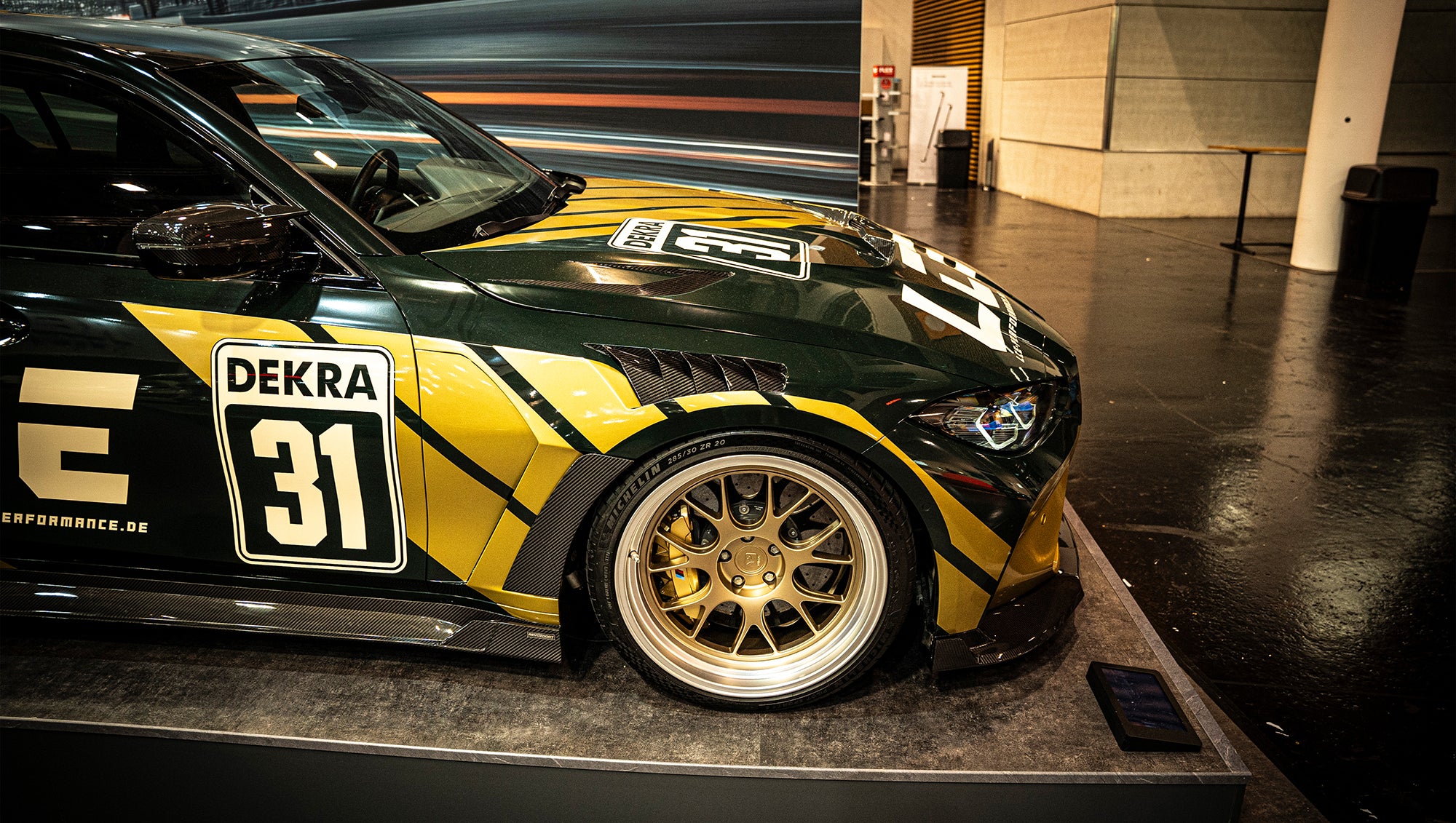 BMW G80 M3 race build with REVOZPORT carbon fiber aero kit, gold wheels, and black-yellow motorsport livery displayed at an indoor automotive exhibition.