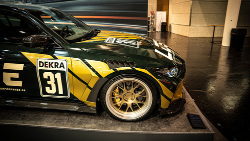 BMW G80 M3 race build with REVOZPORT carbon fiber aero kit, gold wheels, and black-yellow motorsport livery displayed at an indoor automotive exhibition.