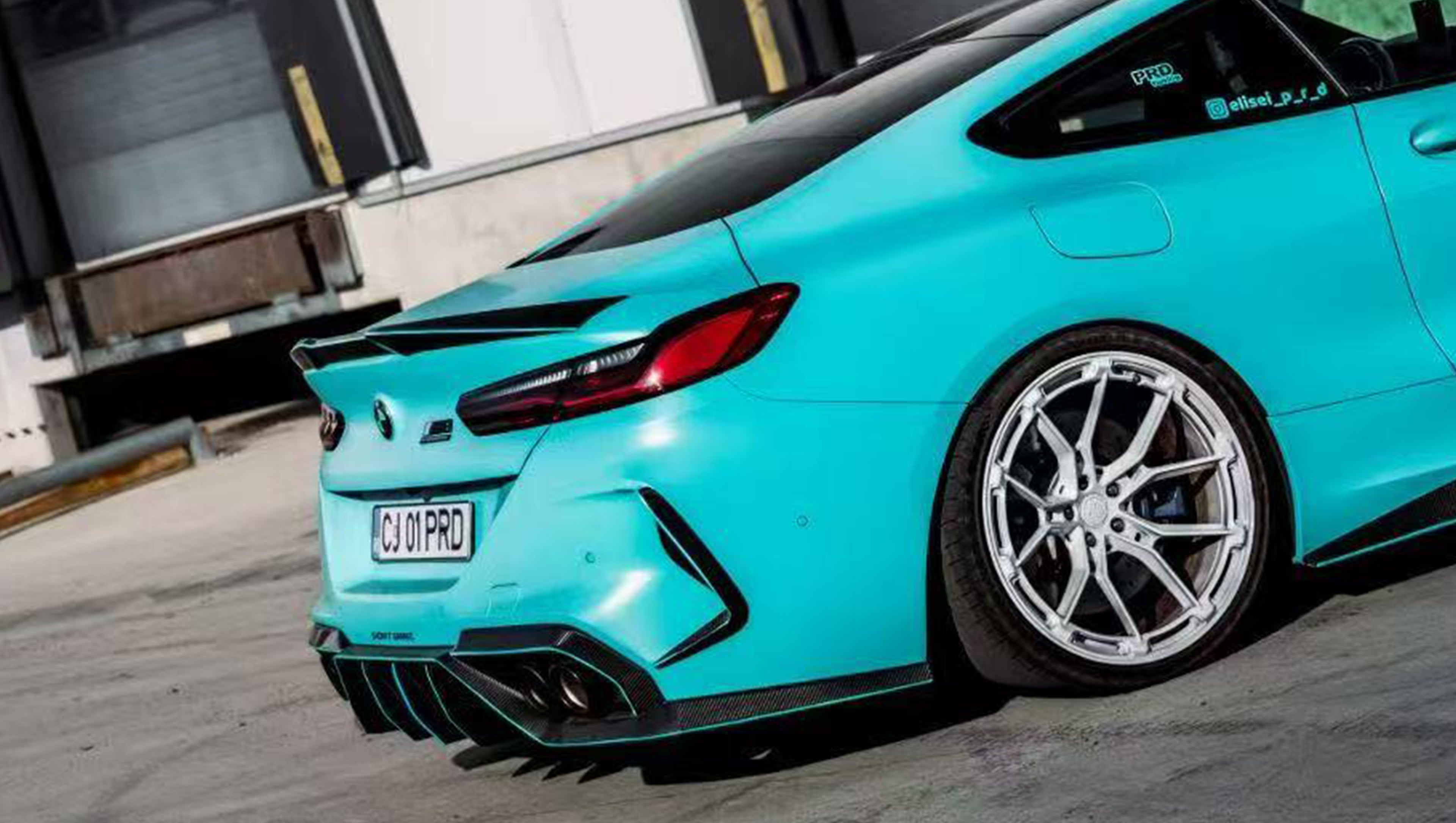 Turquoise BMW F92 M8 featuring a REVOZPORT carbon fiber body kit with front lip, side skirts, black hood accents, side vents, and silver forged wheels parked outside an industrial loading dock.