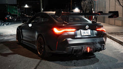 Matte black BMW G82 M4 with carbon fiber front splitter, side skirts, vented hood, bronze wheels, and oversized rear wing parked on a city street at night with headlights on.