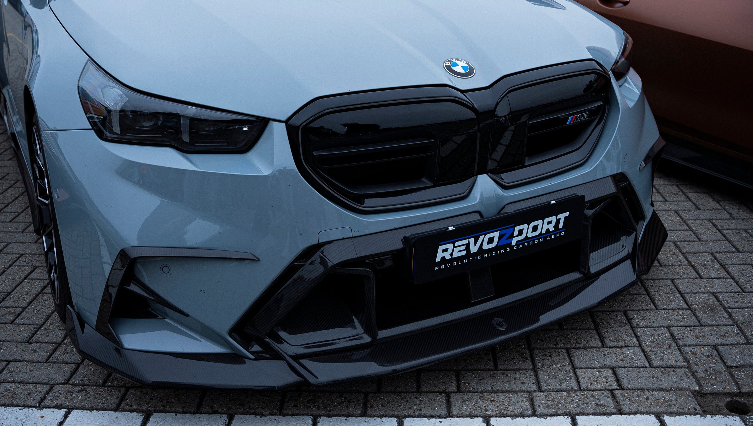 Gray BMW G90 M5 Touring featuring a REVOZPORT carbon fiber front lip, aggressive front aero styling, and black grille displayed at a car event.