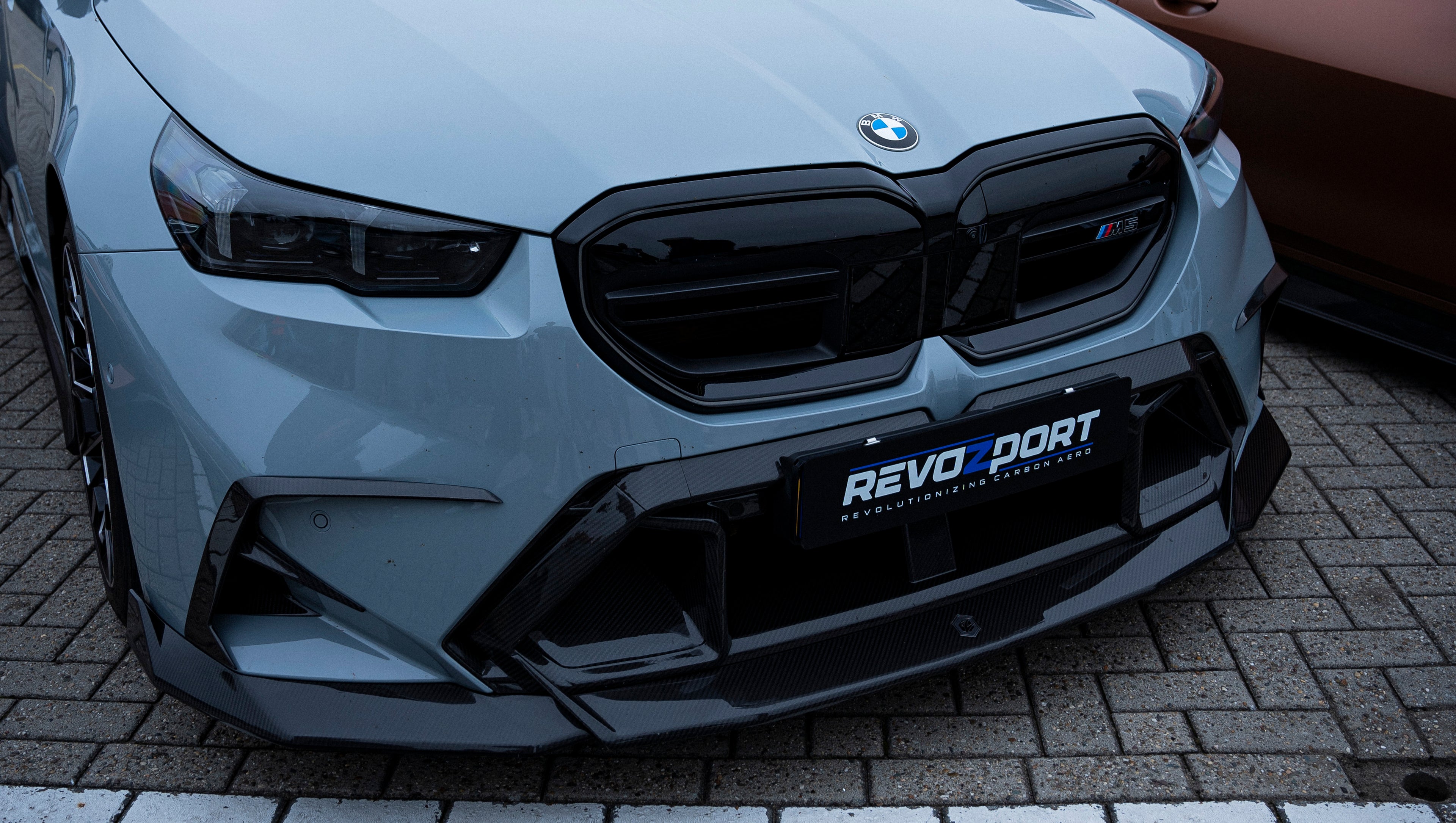 Gray BMW G90 M5 Touring featuring a REVOZPORT carbon fiber front lip, aggressive front aero styling, and black grille displayed at a car event.