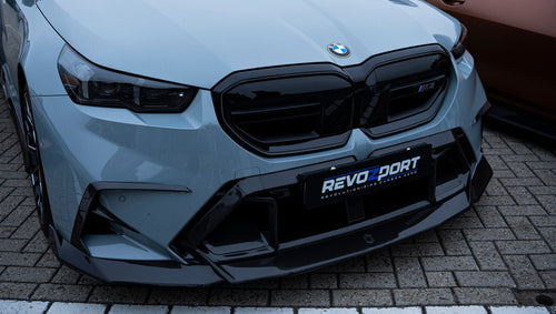 Gray BMW G90 M5 Touring featuring a REVOZPORT carbon fiber front lip, aggressive front aero styling, and black grille displayed at a car event.