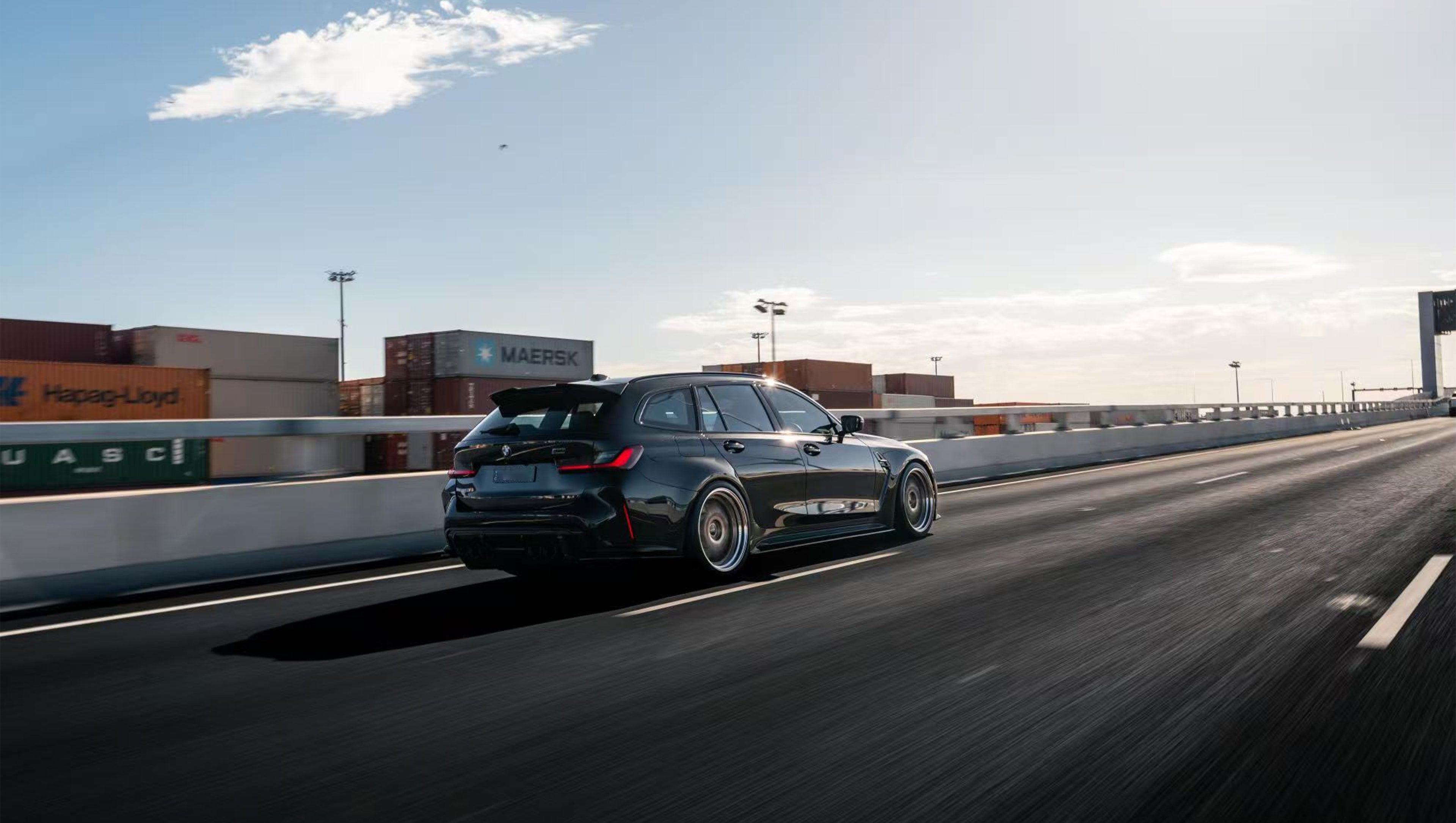 Black BMW G81 M3 Touring featuring a REVOZPORT carbon fiber body kit with vented fenders, side skirts, bronze wheels, and red performance brakes parked beside a stone wall.