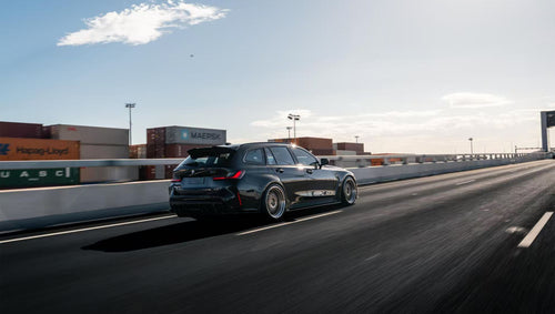 Black BMW G81 M3 Touring featuring a REVOZPORT carbon fiber body kit with vented fenders, side skirts, bronze wheels, and red performance brakes parked beside a stone wall.