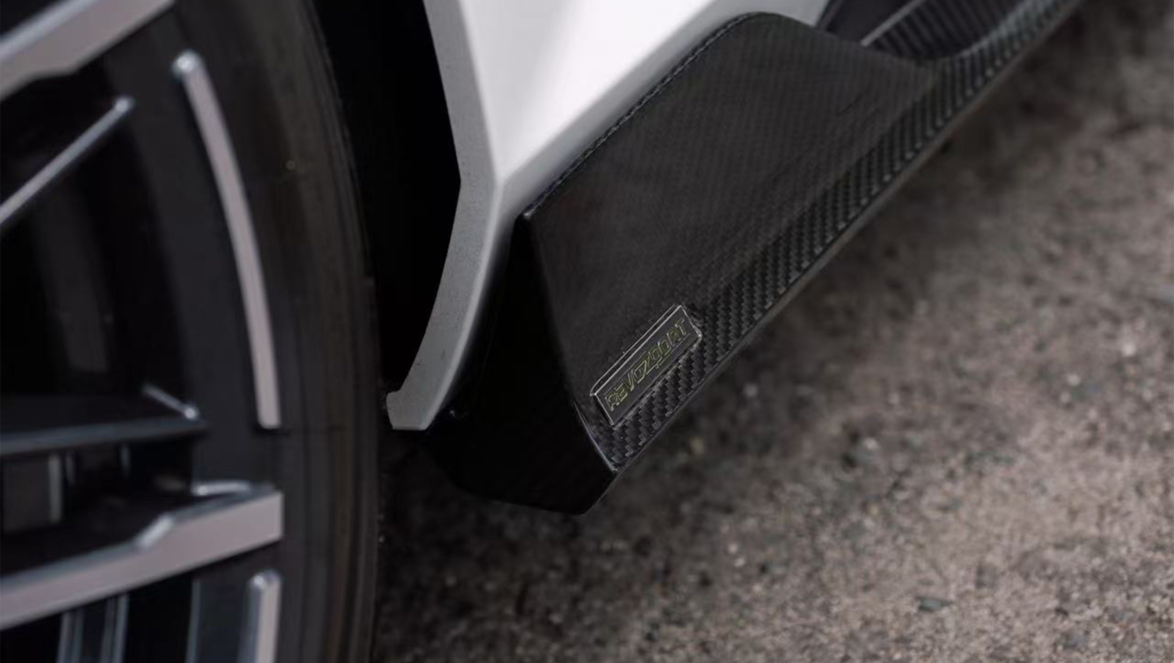 White BMW G90 M5 featuring a REVOZPORT carbon fiber front lip, side skirts, and aggressive front aero styling parked in a natural outdoor setting.