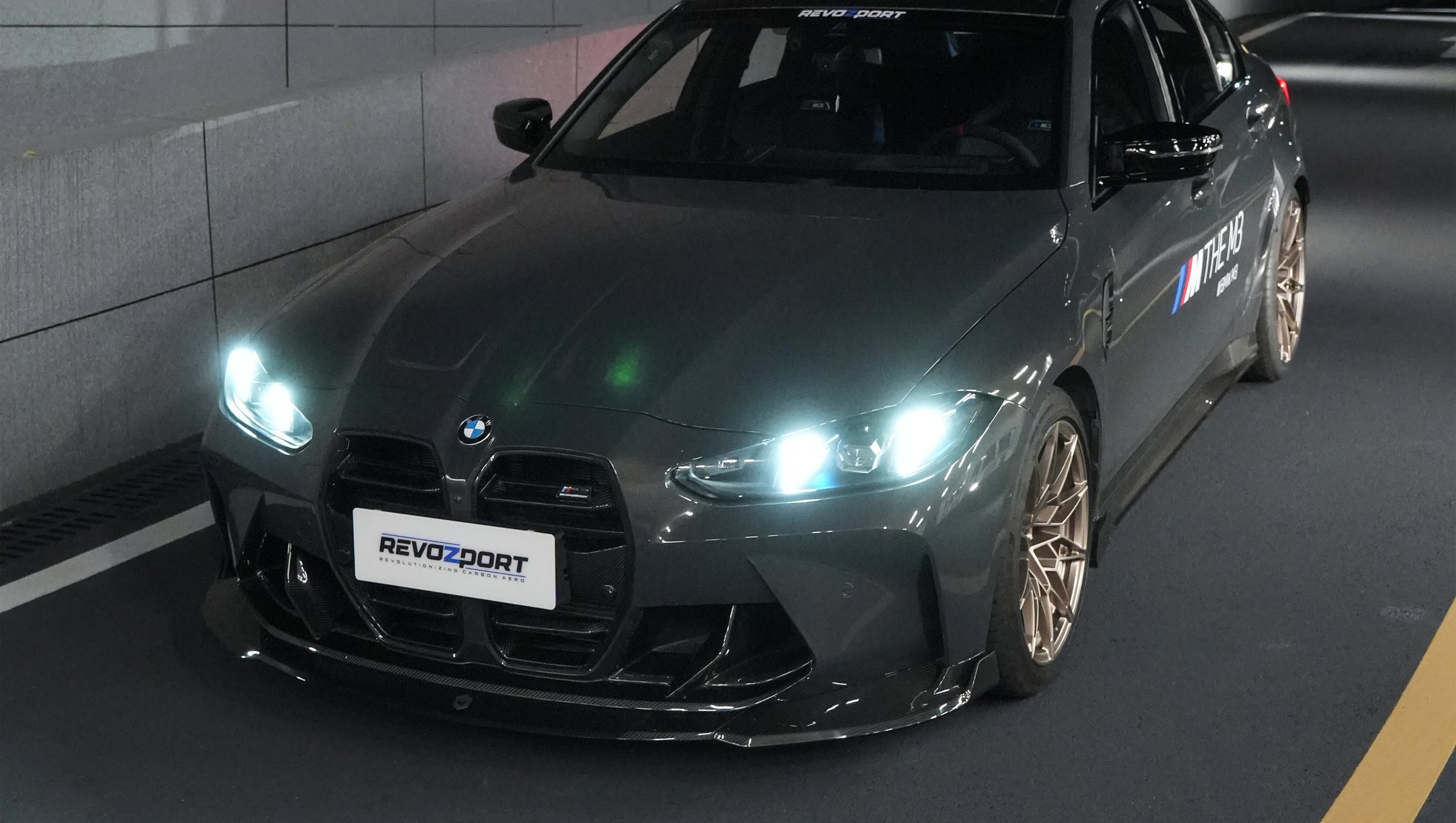 Gray BMW G80 M3 featuring a REVOZPORT carbon fiber front lip, side skirts, bronze wheels, and M The M3 side graphics driving through a tunnel at night.