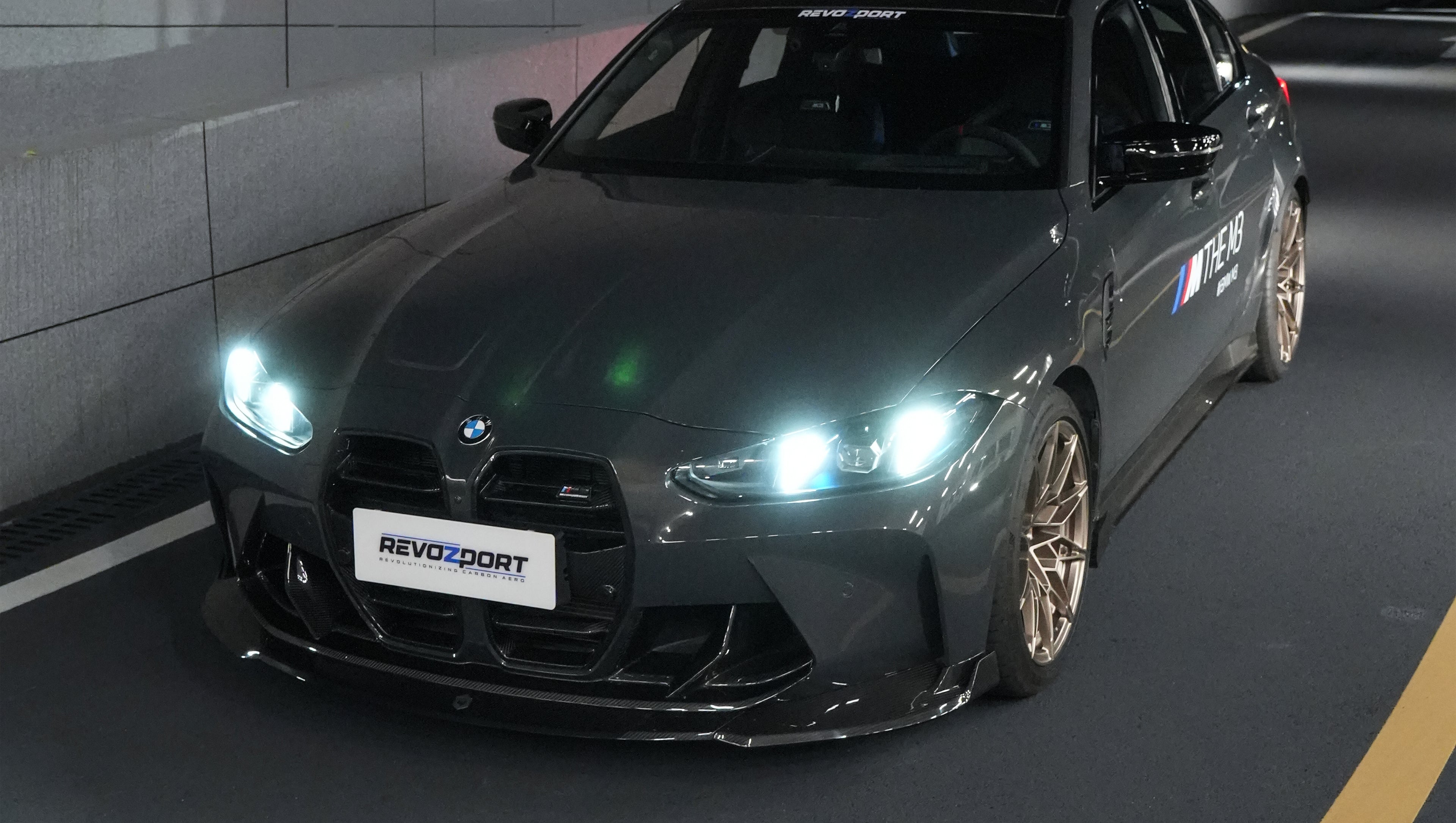 Gray BMW G80 M3 featuring a REVOZPORT carbon fiber front lip, side skirts, bronze wheels, and M The M3 side graphics driving through a tunnel at night.