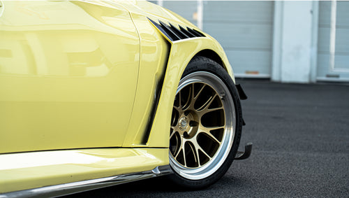 Pastel yellow BMW G82 M4 with carbon fiber front splitter, hood vents, side canards, bronze wheels, and large rear wing parked at a race track facility during golden hour.
