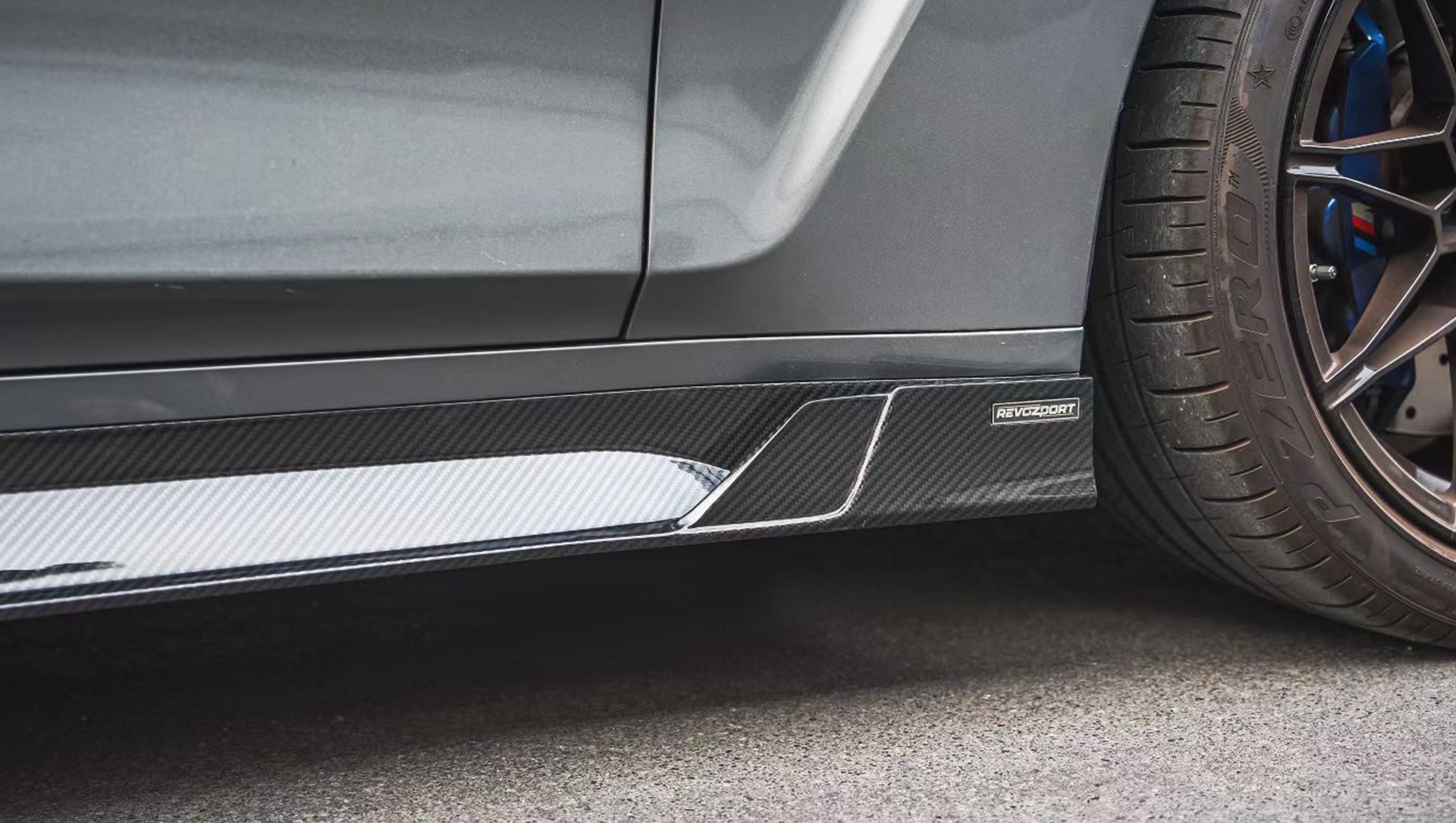 Gray BMW G80 M3 featuring a REVOZPORT carbon fiber front lip, side skirts, and aggressive aero styling parked outside a modern urban garage.